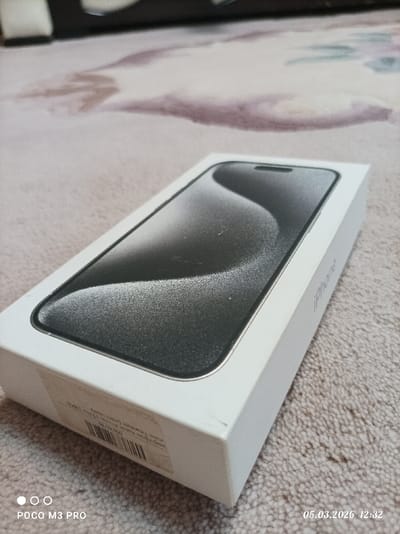 An upward-angled, close-range photograph depicts a white iPhone retail box resting on a light-colored, patterned carpet in Dmitrievskaya, Russia. The box's top surface features an image of a dark smartphone screen with a dynamic, wavy gray design. The word "iPhone" is vertically printed in light gray on the right side of the box. A white label with a black barcode and text, including "128Gb," is visible on the lower left side of the box. The bottom left corner of the box exhibits minor scuffing or a crease. The carpet displays a subtle pattern of muted brown and gray shapes. The background is out of focus, showing indistinct indoor elements. The image bears a timestamp of "05.03.2026 12:32" and "POCO M3 PRO" in the bottom corners. No individuals are present, and no actions or interactions are depicted.