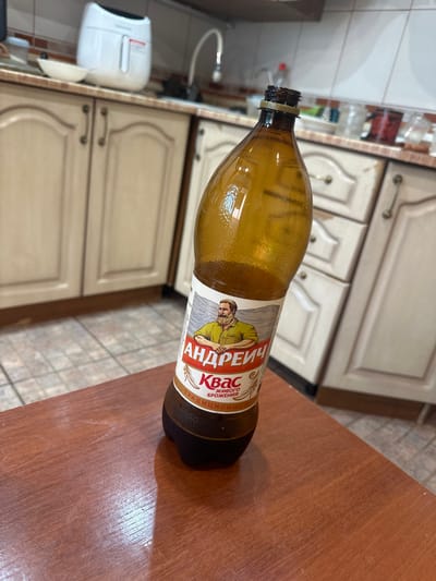 The image shows a close-up of a plastic bottle of "Andreich Kvas" on a wooden table. The bottle is amber-colored and partially filled with a dark liquid. A label on the bottle features a drawing of a man with a beard wearing a green shirt, along with text in Russian. In the background, a kitchen area is visible with cabinets, countertops, and a sink. There is no indication of the specific location in Topki, Russia.