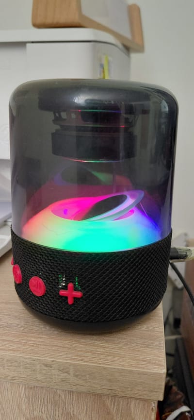 The image shows a cylindrical speaker with a clear plastic exterior, revealing colorful LED lights that cycle through various hues of pink, green, and blue. The speaker has a black mesh fabric covering on its right side, where several red buttons are visible, including a plus-shaped button and other circular buttons. The speaker is placed on a light-colored wooden surface, possibly a shelf or table, and is positioned next to a white appliance. In the background, a portion of a white wall and a wooden object with a curved edge are visible. The lighting suggests it is indoors, likely during the daytime. The scene appears to be a domestic or office setting. There is no visible text in the image.