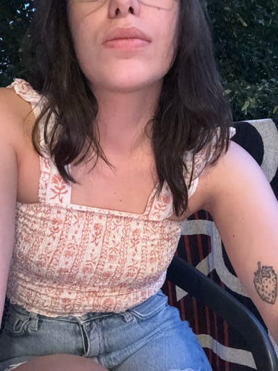 This media file shows a close-up, top-down view of a person from the chest up, lying on their side. The subject has dark hair that falls across their face and chest. They are wearing a light-colored, patterned top with a floral or paisley design in shades of pink and white, and denim shorts. A portion of their arm is visible, showing a tattoo of a strawberry on their upper arm. The background is out of focus, but appears to be an outdoor setting with greenery and what looks like a chair with a striped cushion. The lighting suggests it might be evening. The person's face is partially visible, with lips and part of their nose in the frame, and they appear relaxed. There is no visible text in the media file. The location context provided is El Escorial, Spain.