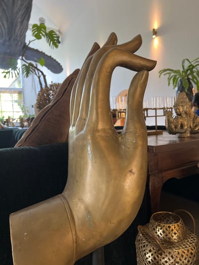 The image displays a large, golden-colored statue of a hand in a pose associated with blessing or peace, a detail likely connected to the cultural significance in Petulu, Indonesia. The hand rests upon a dark-colored surface, which could be a cushion or part of a piece of furniture, with hints of a brown fabric nearby. In the background, a blurred interior setting is visible, featuring plants, a window suggesting natural light, and other decorative objects like a metallic lantern and a smaller statue. The overall scene suggests an indoor setting, potentially a place of worship or a personal shrine, where the statue serves as a central element.