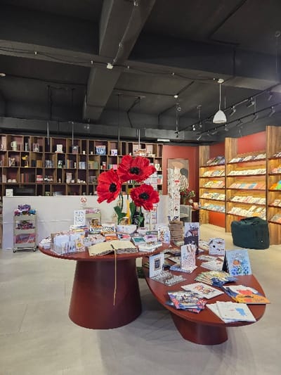 The image depicts the interior of a retail space, specifically a bookstore or gift shop, devoid of people. The ceiling is dark, with exposed ventilation ducts, electrical conduits, and track lighting fixtures, alongside some pendant lights. The floor is light grey. In the midground, two connected, multi-level display tables, made of dark reddish-brown wood, are laden with various merchandise including books, greeting cards, and notebooks. Three prominent, large artificial red poppy flowers are centrally displayed on the taller table. To the left background, a tall, dark wooden bookshelf unit with numerous cubbies holds books and decorative items. Adjacent to this, a white service counter features a "No Smoking" sign, and a metal rolling cart with multiple shelves displays colorful items. To the right background, more wooden shelving units present books and magazines face-out. A dark green fabric bag rests on the floor near these shelves. A framed artwork depicting a figure in a striped garment is visible on a dark red wall section between the central display and the right-hand shelving. The specific location within the city of Bogorodica, North Macedonia, cannot be determined from the visual information provided.