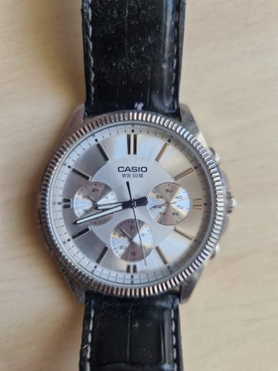 This is a close-up, eye-level, horizontal shot of a silver Casio wristwatch with a black leather band. The watch face is silver with silver hour markers and three sub-dials. The main dial shows "CASIO" and "WR 50M". One sub-dial is labeled "SU MO TU WE", indicating a day of the week function. Another sub-dial shows numbers "20" through "24", possibly for a 24-hour display. The third sub-dial shows numbers "15" through "31", likely for the date. The watch has a ribbed bezel and two pushers on the right side. The black leather band has white stitching. The background is a light-colored, textured surface, possibly wood or a table. The lighting suggests it is daytime. There are no people visible in the image, and no specific location cues for Rikava, Latvia, are present beyond the provided context.