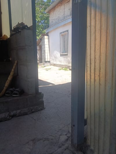 This image captures an outdoor scene in Ananyevo, Kyrgyzstan, likely during daylight hours given the bright sunlight casting shadows. The setting appears to be a utilitarian, possibly residential, area with a mix of construction materials and simple structures.
In the foreground, to the left, is a concrete structure with a rough finish. Resting on it is a pair of dark-colored sandals, suggesting recent use or current accessibility. Leaning against this structure is a broom with a woven straw head, another indicator of daily chores or maintenance. Further back and to the left, a white corrugated metal wall is visible, partially obscured by shadows.
The middle ground opens into a courtyard or pathway. The ground is paved with cracked concrete or asphalt. To the right of the pathway is a building with white stucco walls and a light-colored tiled roof. A single window with a light frame is visible on this building. Next to it, a smaller, rougher structure made of concrete blocks with a wooden door stands, and a shovel with a dark blade is propped against it. Green vegetation, likely weeds or small plants, grows sparsely in patches on the ground and around the base of the structures.
On the right side of the image, a large, vertical, corrugated metal structure, possibly a gate or part of a shed, dominates the frame. It has a yellowish-brown hue, suggesting it might be made of aged metal or plastic, and is illuminated by direct sunlight. The texture is distinctly ribbed.
The overall impression is one of a lived-in, functional space, perhaps a backyard or an alleyway. The sunlight suggests a warm day, and the general appearance of the area is modest and perhaps a bit weathered. There is no text visible in the image. The scene conveys a sense of ordinary life in a rural or semi-urban setting.
