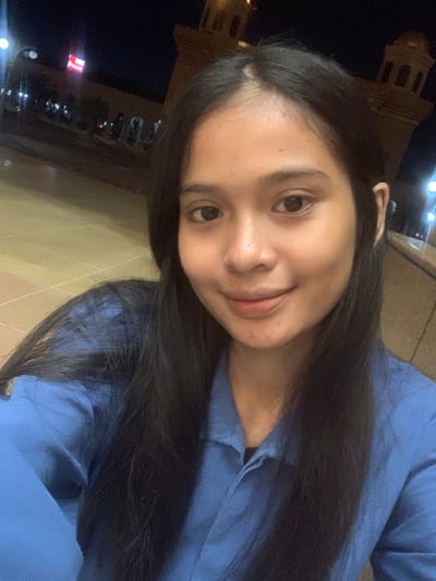 The image is a selfie-style portrait of a young woman taken at night, rotated clockwise. The primary subject is a young woman with long, dark hair and dark eyes, looking directly at the camera with a subtle, pleasant smile. She is wearing a blue long-sleeved top or shirt.

The background is dark, indicating nighttime, with various artificial lights visible in the distance. To the upper left, a faintly illuminated building with distinct architectural features, possibly a tower or a prominent public structure, is visible against the dark sky. Below the woman, a paved ground surface is visible. Additional crops reveal close-ups of her face, hair, and the blue garment, as well as more details of the dark, urban background with scattered bright lights and what appears to be a red and white sign or object in the distance. The scene suggests an outdoor setting, likely a public area or street in Samarinda, Indonesia. No discernible text is visible.