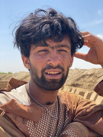 A medium shot captures a man in Deh-E Bagh, Afghanistan. He has dark, curly hair and a beard, with a silver chain around his neck. He wears a brown, patterned shirt over a white undershirt. His right hand is near his head. In the background are piles of earth or bricks and a clear, blue sky.