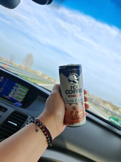 The image displays a close-up, first-person perspective from within a vehicle, located in Manolsko Konare, Bulgaria, looking out through the windshield during daylight hours. A human right hand, adorned with multiple bracelets (including one black and red woven bracelet with a silver heart charm, and another composed of dark beads), occupies the center-right of the frame, holding an upright silver beverage can. The can is labeled "ICE COFFEE Double Espresso" with "+20% CAFFEINE" and "WITH REAL COFFEE EXTRACT," featuring a stylized white animal head logo (resembling a wolf or fox). To the left, a portion of the vehicle's dark dashboard is visible, including a navigation screen displaying a map interface with colored routes. The background, viewed through the windshield, shows a multi-lane highway or road with safety barriers and green roadside foliage. The sky is clear blue with sparse white clouds, and distant trees with bare branches are visible on the horizon.