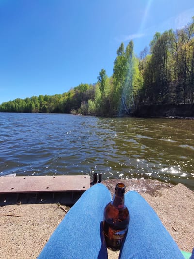 A point-of-view shot shows a person wearing blue jeans sitting by a body of water under a clear blue sky. The water appears to be part of a lake or river. Green trees line the opposite shore, indicating a natural or park-like setting. A brown glass bottle sits on the person's lap between their legs. The location is likely near Икское Устье, Russia, based on geographical context. The setting is outdoors, with the person positioned on what appears to be a concrete or stone structure next to the water. The scene suggests a relaxed or contemplative moment.