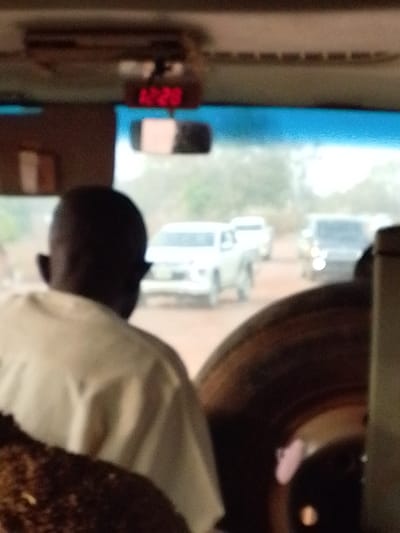 The image, captured from inside a vehicle in Oturkpo, Nigeria, depicts an exterior scene through the front windshield. A digital clock displaying "12:20" is mounted centrally above the windshield, with a rearview mirror positioned below it. The back of an individual's head and shoulders are visible in the immediate foreground, showing short, dark hair and a light-colored garment. To the lower right, a dark, circular object, possibly a spare tire, is partially visible. Outside, on an unpaved dirt road, traffic is present. A white pickup truck is prominently featured, facing away from the camera, with a lighter-colored vehicle behind it. Further ahead, a dark-colored vehicle is visible, potentially facing the camera. The background indicates a hazy outdoor environment with sparse distant vegetation under daylight conditions. No specific actions or interactions are discernible.