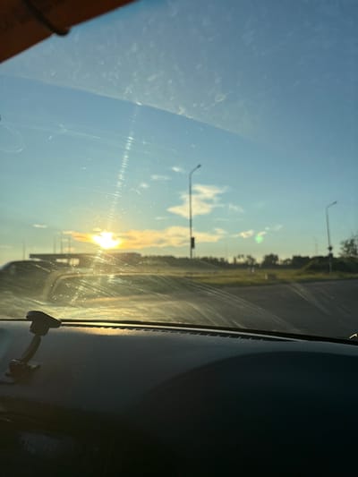The image depicts a view from inside a vehicle looking through the windshield at a sunset. The sun is low on the horizon, casting a warm glow across the sky, which is a mix of blue and yellow hues. Clouds are present, with some illuminated by the setting sun. In the distance, there appears to be a road or highway, with streetlights visible on either side. A structure, possibly a gas station or a building with tall poles, is also in the background. The dashboard of the car is visible in the foreground, with a black textured surface and a mount for a device on the left side. The windshield has visible streaks and smudges. The overall environment suggests an outdoor, likely rural or suburban setting, potentially on the outskirts of a city or town in Вяйнянскі сельскі Савет, Belarus.
