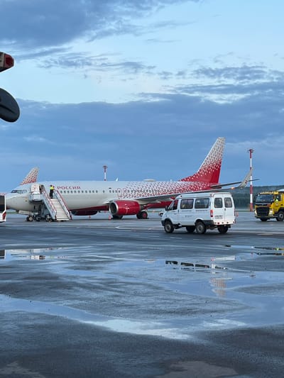 A vertical, medium shot captures an airplane at an airport, likely Yemelyanovo Airport in Russia, based on the visible text. The airplane, predominantly white with red accents and a distinctive red and white patterned tail fin, is parked on a wet tarmac. A set of mobile stairs is connected to the aircraft's passenger door, with a person in a yellow vest visible on the stairs, suggesting boarding or disembarking.
Beside the airplane is a white minibus with multiple windows and four wheels. Text on the side of the minibus is partially visible, appearing to be in Cyrillic script. Further in the background, a yellow service vehicle with hazard lights is parked on the tarmac. The sky above is overcast with visible clouds, indicating a cloudy day, and the time of day appears to be daytime. The wet tarmac suggests recent rain or damp conditions. The overall scene depicts a typical airport operation. The visible text on the airplane reads "РОССИЯ", which translates to "RUSSIA."