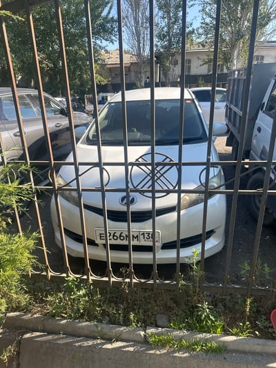 The image shows a white car parked behind a metal fence, viewed from a low angle. The car has a license plate that reads "E266NM 138 RUS". A circular metal decoration with intersecting lines is placed on the hood of the car. In the background, there are trees, buildings, and other vehicles, including a truck to the right. The scene appears to be outdoors, likely in a residential or commercial area. The overall impression is of an ordinary street scene, possibly in the city of Kara-Suu, Kyrgyzstan, given the Russian license plate and the presence of buildings and parked vehicles typical of an urban environment.