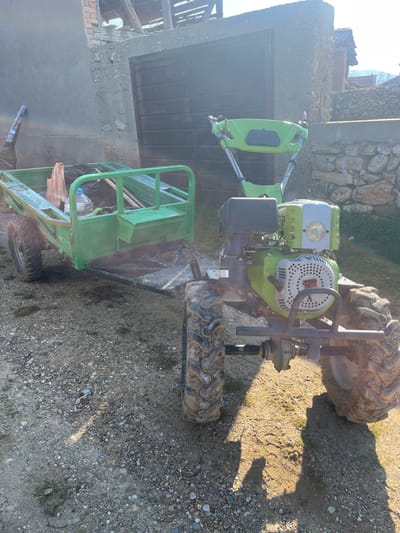 A light green walk-behind tractor (motocultor) is stationary on a gravel and dirt surface, facing left, in Smochevo, Bulgaria. A two-wheeled, light green utility trailer is hitched to the rear of the tractor. The tractor features a prominent engine block with a visible "13 HP" label, a silver-colored front grille, and a circular headlight. Its deeply treaded tires are coated with dried mud. The trailer's open bed contains a clear plastic water bottle, a white plastic shopping bag, and several long-handled tools, likely garden implements. In the background, a building with a partial concrete facade, dark brown wooden double doors, and exposed wooden roof rafters is visible. To the right, a dry-stacked stone wall is present. The scene is brightly illuminated by direct sunlight, creating noticeable lens flare and casting distinct shadows on the ground. No individuals are present.