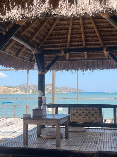 The image shows a view from under a thatched-roof gazebo, looking out over the ocean. In the foreground, there is a rustic wooden table and a bamboo floor. On the table, there is an ashtray, a box of tissues, and a glass jar filled with straws. In the middle ground, there is a railing overlooking the water, with a wooden bench on the right. The water is a clear turquoise color and there are several boats visible, including a small blue boat in the distance and a sailboat further out at sea. In the background, there are hills and mountains. The overall scene appears to be a peaceful and relaxing beachfront setting.