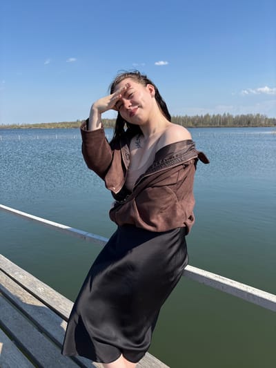 A woman is posing on a wooden pier overlooking a body of water, possibly in Crokhovo, Russia, given the location specified. She is wearing a black dress and a brown suede jacket. Her hair is dark, and she has a tattoo on her upper chest. The woman's right hand is raised towards her face, possibly shielding her eyes from the sun. The pier has a white railing. The background includes a blue sky with some clouds and a distant shoreline with trees.