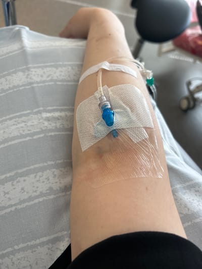 The image shows an arm with an IV drip in it, resting on a light gray and white striped blanket. The IV line is secured to the skin with medical tape and a transparent dressing. The person's skin is fair. The background is blurred but suggests a medical setting, possibly a hospital room, with a dark chair and some medical equipment visible. The location of Helsingborg, Sweden is not ascertainable from the visual information presented in the image.
