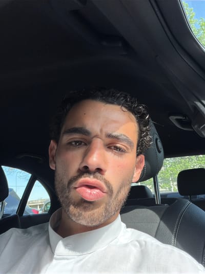 A selfie shows a man with dark curly hair, light skin, and a trimmed beard inside a car. He is wearing a white shirt. The interior of the car is dark, with black leather seats and trim. Outside the car window, blurry trees and buildings are visible. The location cannot be determined to be Mont-de-Marsan, France from the given image alone.