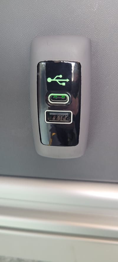 A close-up shot of a modern power outlet with two USB ports is visible. The outlet is a dark, reflective material, likely plastic, with a lighter gray casing. A green USB symbol is illuminated above the ports. The top port is a USB-C port, also illuminated with a green light. The bottom port is a standard USB-A port. The outlet is mounted on a textured gray surface, possibly a wall or seat in a vehicle, which is characteristic of public transportation or a modern office environment. A blurred metallic bar runs horizontally across the lower part of the image, suggesting the presence of other furniture or fixtures. The image is taken from a slightly downward angle. The location is not within the city of Obertshausen, Germany.