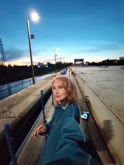 In Sheksna, Russia, a woman with blonde hair and red nail polish stands on a concrete embankment at dusk. She is wearing a blue denim jacket and has her left hand resting on the railing. A smartphone is placed on the ground near her right leg. The embankment runs alongside a body of water. In the background, a tall metal tower and several streetlights are visible, along with silhouetted trees and buildings. The sky is a gradient of blue and orange, indicating the setting sun.