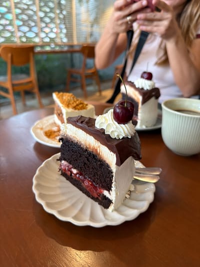 The image depicts an indoor dining scene, likely a cafe, in Chiang Dao, Thailand, focusing on three slices of cake and a cup of coffee on a wooden table. In the immediate foreground, a slice of multi-layered cake rests on a white scallop-edged ceramic plate. This cake features a dark brown chocolate base, a red translucent fruit filling (likely cherry), white cream or frosting layers, and a top layer of dark chocolate ganache that extends over the sides. It is garnished with a dollop of white whipped cream and a single red cherry with a green stem. A silver fork is partially visible on the plate next to this cake.

To the right, another slice of chocolate cake on an identical white plate is visible, structured similarly with dark cake, white cream, chocolate ganache, whipped cream, and a red cherry with a stem. Further to the left and rear, a third slice of cake, also on a white plate, presents a different appearance, with white frosting and a covering of light brown crumbs. A light green, ribbed ceramic mug containing a dark liquid, presumably coffee, is positioned to the right of the second cake.

The table surface is made of polished reddish-brown wood, exhibiting minor scratches and reflections. In the blurred background, a person, identified as female by visible features including long light hair, is holding a pink smartphone with both hands, angled towards the table as if capturing a photograph or video. She wears a light-colored top and a dark strap, possibly from a bag, across her shoulder. A silver-colored ring is visible on her left ring finger. Behind the person and the table, out-of-focus wooden chairs and tables are discernible, along with horizontal window blinds. Through the blinds, a blurred outdoor environment with green foliage and abstract colorful light reflections is visible.