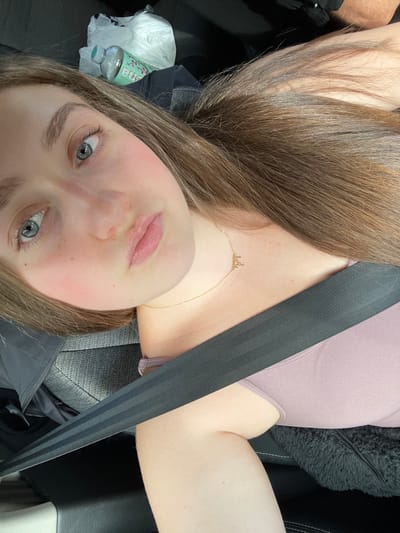 A woman in a car takes a selfie while wearing a mauve top and a seatbelt. She has long brown hair and a gold necklace. A water bottle and a plastic bag sit on the car seat next to her. She has fair skin and blue eyes. The car seat is dark gray leather.