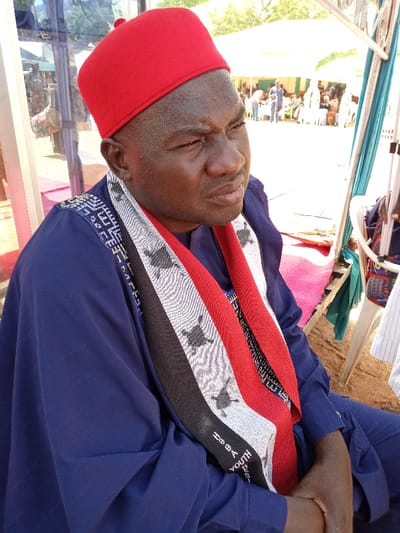 The image shows a man wearing a red cap, a navy blue long-sleeved shirt, and a white, black, and red patterned scarf. The scarf reads "Youth, Be a..." The man is sitting in a chair. He is looking to his right. He appears to be in his forties, has dark skin, and has short black hair. In the background, there are several people. It appears to be a sunny day. The background is a blur, making it difficult to discern any further details. There are large tents visible in the distance. This image was taken in Zum, Nigeria, at the coordinates 10.116 latitude and 13.046 longitude.