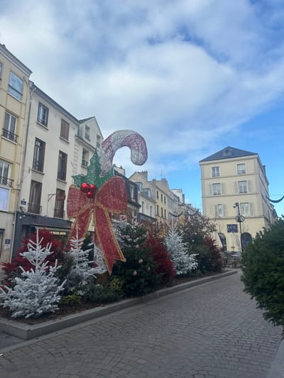 The image displays a Christmas-decorated urban street in Saint-Germain-en-Laye, France, under an overcast sky. In the foreground, a large, illuminated decoration features a white and red striped candy cane, green holly leaves with red berries, and a prominent red glittery bow. This decoration is situated within a raised planter bed that extends along the left side of a cobblestone pathway. The planter bed contains a dense arrangement of various small Christmas trees, including bright red, heavily frosted white, and green evergreens, some adorned with red and white striped baubles. The cobblestone street is empty. Along the left, a row of multi-story buildings with light-colored facades and multiple windows lines the street; a shopfront labeled "LE REVEIL MATIN" with "BAR" above it is visible. Further down the street on the right, a pale yellow four-story building with a dark mansard roof and shuttered windows stands, with an "LCL" sign and outdoor seating visible below. Overhead Christmas garlands are present further down the street. The sky is light blue with scattered white clouds.