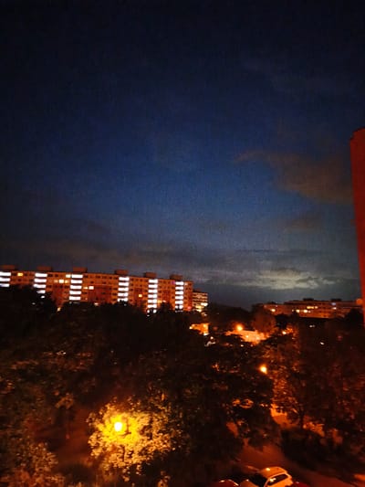 The image captures a night scene in the city of Most, Czechia. A large apartment building with numerous illuminated windows dominates the mid-ground, stretching horizontally across the frame. In the foreground, dark silhouettes of trees are visible, with a bright, orange streetlamp casting a warm glow on the foliage. The sky above is a deep, dark blue, with some faint clouds and distant city lights visible on the horizon. A red building is partially visible on the far right edge of the image. A white car is parked in the lower right corner of the frame. The overall impression is one of a typical residential area at night.