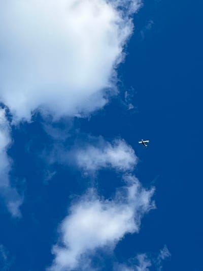 The image shows a bright blue sky with scattered white clouds. A small white and blue airplane is visible in the upper right quadrant of the image, flying against the sky. The airplane is viewed from below. The location is not specified in the image.