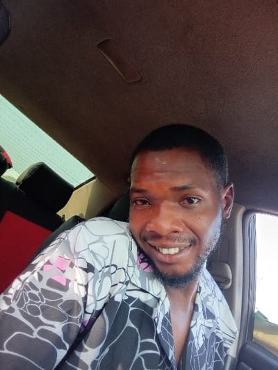 A smiling man with a beard, wearing a patterned gray and black shirt with pink accents, is angled to the left in the frame. His eyes are open and directed at the viewer, showing his teeth. The photo appears to be taken from inside a vehicle, with the interior ceiling behind him. A car window and part of a seat are visible in the background.