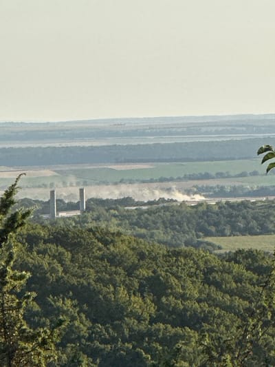 The image shows a panoramic view of a rural landscape, likely in or around Pryvorottia Druhe, Ukraine. In the foreground, a dense forest of green trees dominates the lower half of the frame. Beyond the forest, the middle ground features a more open area with agricultural fields and what appear to be two tall, slender structures, possibly chimneys or industrial towers, situated between patches of trees. A plume of dust or smoke is visible in the distance, suggesting some activity such as farming or construction. The background consists of layered, muted green and blue hills fading into a hazy, pale sky. The overall impression is of a serene, expansive countryside.