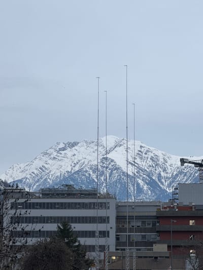 This image depicts an urban landscape in Visp, Switzerland, under an overcast sky, with snow-capped mountains in the background.

The scene features a dominant, multi-story, light-grey building with multiple rows of rectangular windows across its facade, located in the left-center midground. The rooftop of this building is partially obscured by scaffolding or construction materials. To the right of this structure, a multi-story building with a reddish-brown facade is visible, exhibiting illuminated windows on its upper floors. Further to the right, a black and yellow tower crane is partially visible.

Four tall, slender, vertical metallic masts or antennas rise from the midground, extending significantly above the building rooftops and partially obscuring the mountain range. Each mast features a small horizontal element at its apex.

In the background, large, rugged mountains are heavily covered in white snow, indicating a winter or early spring setting. The sky is uniformly light grey and overcast. The immediate foreground contains leafless trees and shrubs, partially obscuring the lower sections of the buildings. A single evergreen conifer tree is situated between the central buildings. No individuals or direct human actions are observable within the frame.