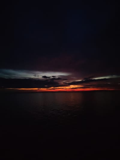 A twilight scene over the sea is depicted, with the sky dominating the upper portion of the frame. The sky transitions from a deep, dark indigo at the top to hues of orange and red near the horizon, indicating a sunset or sunrise. Clouds are present, varying in color from dark gray to lighter shades with hints of pink and yellow. Below the sky lies the sea, which appears calm and dark, reflecting the muted colors of the sky. The horizon line is distinct, separating the sky and the water. There are no visible people, animals, or man-made structures in the image. The overall atmosphere is serene and subdued, characterized by low light conditions. The scene is likely captured in a coastal area.
