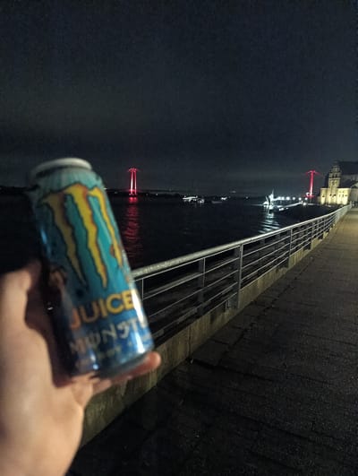 A hand holds a blue Monster Juice can in the foreground. The background features the Rhine River at night, with the Rhine Bridge illuminated in red light. Along the right edge is a walkway with a metal railing. In the distance to the right of the bridge is the cityscape of Emmerich on the Rhine, Germany. Several lights reflecting off the water are visible. The sky is dark and overcast.