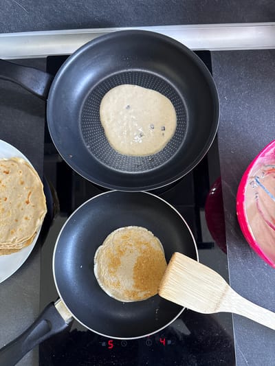 In Skravena, Bulgaria, a close-up, top-down view captures a kitchen scene where pancakes are being prepared. Two non-stick frying pans are situated on a black induction cooktop. The upper pan contains a thin layer of batter with small dark specks, indicating it's in the early stages of cooking. The lower pan holds a partially cooked pancake, golden brown on one side and displaying a textured surface. To the left, a white plate is stacked with several finished, golden-brown pancakes. To the right, a bright pink bowl holds pancake batter, with a wooden spatula resting on its edge. The induction cooktop displays digital readouts of "5" and "4" on its respective zones. The environment is a kitchen counter, with dark gray speckled material visible in the background.