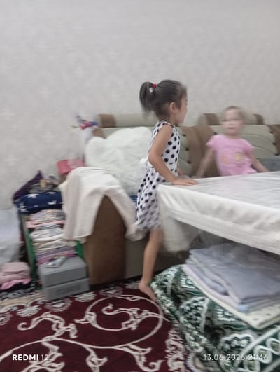 In a domestic indoor setting, two young girls are visible, one wearing a black and white polka dot dress and the other a pink t-shirt. The girl in the polka dot dress is standing and appears to be interacting with the other girl who is seated on a sofa. The environment includes a sofa, a stacked collection of folded fabrics and garments, and a rug with a red and white patterned design. A timestamp in the lower right corner indicates "13.06.2026 21:46," and "REDMI 12" is in the lower left corner, suggesting the image was taken with a Redmi 12 phone. While the image context suggests a residential area, specific details about the city of Bekabad, Uzbekistan are not discernible from the visual information provided.