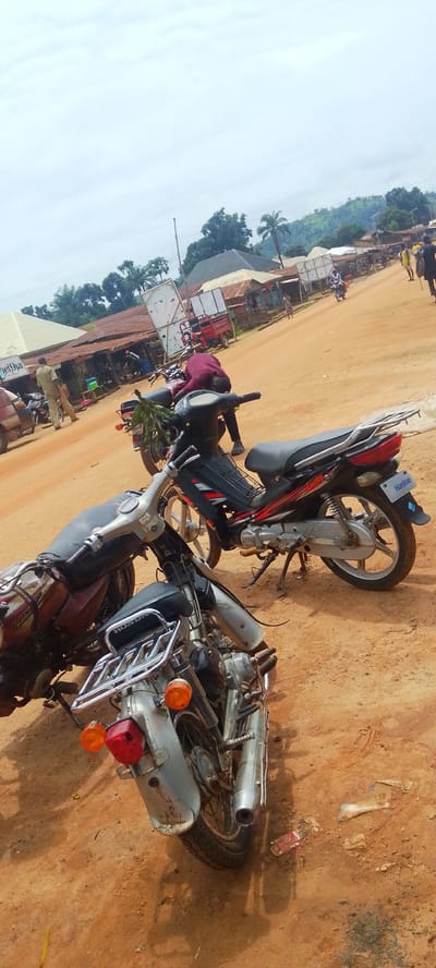 The image depicts a street scene in Baissa, Nigeria. The foreground is dominated by two parked motorcycles, one a silver and black model with a chrome luggage rack and the other a darker, possibly maroon, motorcycle partly obscured. In the middle ground, a person in a dark maroon shirt is bending over a motorcycle, seemingly working on it. To the left of this person, another motorcycle, black with red accents, is parked. Further in the background, a dirt road stretches into the distance, lined with modest buildings, some appearing to be shops with corrugated metal roofs. Several people are visible along the road, some walking and others near the buildings. One person in a light-colored shirt stands near a building on the left. Another person in a yellow shirt is further down the road, carrying a bag. A red cart, possibly a type of delivery vehicle, is parked near one of the buildings. The overall scene suggests a busy, possibly market day, in a rural or semi-urban Nigerian setting. The sky is overcast, indicating a cloudy day. The terrain appears somewhat hilly in the far background, with lush green vegetation. Visible text includes "Lucky" and "Haolue" on the black and red motorcycle.