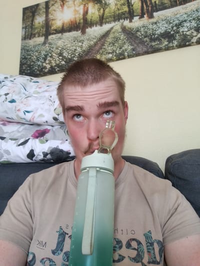 A young man with light brown hair and stubble is holding a large, translucent green water bottle with a straw to his face, as if to drink. He is looking upwards, with a thoughtful or playful expression. He is wearing a light tan t-shirt with a graphic print on the front. The t-shirt has the text "WM" on the left side, and on the right, it appears to have some stylized text that is partially obscured and reversed, possibly "out loud" or similar.
Behind him, a stack of pillows with a floral pattern is visible. Further in the background, a large canvas artwork depicts a sun-dappled forest path lined with white flowers. The scene appears to be indoors, with a pale yellow wall behind the man and a dark gray couch to his right. The lighting suggests it is daytime, possibly late morning or early afternoon, given the bright sunlight depicted in the artwork and the natural light entering the room. The overall mood is casual and relaxed. The location context is Cēsis, Latvia.