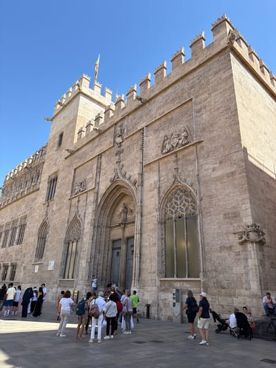 The image depicts the La Lonja de la Seda (Silk Exchange) in Valencia, Spain, under a clear blue sky. The building is constructed of light-colored stone and features intricate Gothic architecture, including pointed arches, ornate carvings, and battlements. A flag, possibly the Valencian flag, flies from a tower.
In the foreground, a paved plaza is populated by a group of people. Some are standing and looking at the building, while others are walking or seated. There are several individuals dressed in casual summer attire. One person is taking a photograph with a DSLR camera. A stroller with two children is visible near the right side of the frame, and a bicycle is parked nearby. The overall scene suggests a tourist attraction on a sunny day. The building itself is a UNESCO World Heritage Site.