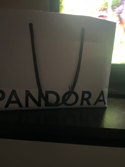 A white Pandora shopping bag with black handles is centered in the frame. The Pandora logo is printed in black on the lower portion of the bag. The bag is placed on a dark surface, possibly a table, in front of a brightly lit screen, which appears to be a television displaying a colorful image. The overall lighting is dim, with the screen providing the primary light source. There are no people or animals visible in the image. The background is out of focus due to the shallow depth of field. The image does not provide any specific geographical information or context that would allow for its placement in Petrevene, Bulgaria.