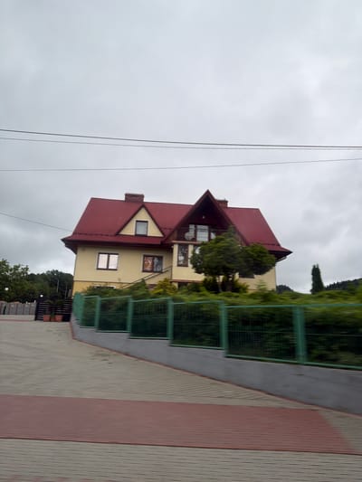In Wilkowisko, Poland, a two-story house with a prominent red, pitched roof dominates the center of the frame. The house is painted a light yellow and features multiple windows, including some with reflective glass. A chimney made of brick is visible on the roof. In front of the house, a green metal fence encloses a landscaped area with various bushes and trees. To the left of the house, a paved walkway with a red accent strip leads away from the viewer. The sky is overcast and gray. Power lines are visible stretching across the upper portion of the image. The overall scene suggests a residential area on a cloudy day.