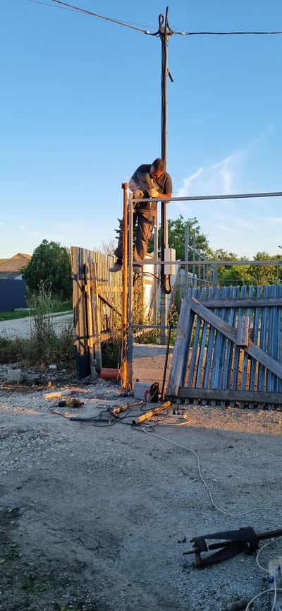A man is welding a metal structure outdoors, possibly in a rural or industrial setting, with a clear blue sky overhead. He is wearing a dark t-shirt and work pants, and appears focused on his task, with sparks flying from the welding. The structure he is working on seems to be a part of a larger framework, with metal poles and beams visible.
In the background, there's a wooden fence and some greenery, suggesting an outdoor location. On the ground near the welding site, several tools are laid out, including a grinder, a welding machine, and some cables, indicating ongoing construction or repair work. The lighting suggests it is either early morning or late afternoon, with warm sunlight casting shadows.
The scene is set in Ленинское сельское поселение, Ukraine. There is no visible text in the image. The overall mood is one of diligent work and industry.