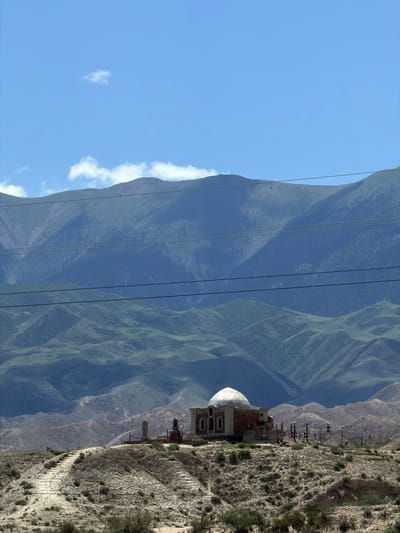 The image shows a mosque with a dome and arched windows on a hillside in the city of Tong, Kyrgyzstan. The mosque is constructed of stone and brick. The surrounding landscape is dry and arid, with sparse vegetation. In the background, there are layered mountains covered in green vegetation. Power lines are visible in the foreground, crossing the frame horizontally. The sky is blue with a few wispy clouds. There are no people visible in the image.