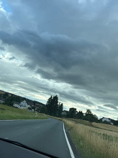 The image depicts a rural roadside view in Oberwetz, Germany. The scene is dominated by a dramatic sky filled with a mix of dark, heavy clouds and lighter, wispy formations, suggesting impending or recent rainfall. The time of day appears to be late afternoon or early evening, given the quality of light.
To the right of the asphalt road, which has a white lane marking, a grassy verge is visible, transitioning into a field of dry, golden grass. Beyond the field, a few houses with red and white roofs are nestled amongst trees. Dark green evergreen trees and some deciduous trees with lush foliage are scattered throughout the landscape. In the distance, two wind turbines are faintly visible, common features in rural German landscapes.
The perspective suggests the photo was taken from inside a vehicle, possibly from the passenger seat, as indicated by the shadowed area in the lower right corner which appears to be part of the car's interior. The overall mood is somewhat somber due to the heavy clouds, yet also serene, characteristic of a peaceful countryside setting. There is no visible text in the image.