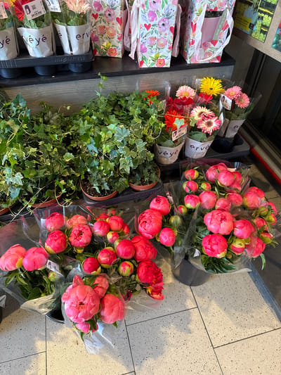 The image shows a display of flowers and plants in a store, likely in Riedlingen, Germany, indicated by the "ervé" and "véyé" packaging. The floral arrangements include bouquets of pink peonies and gerberas in various colors (pink, yellow, red, and orange). There are also potted ivy plants. Floral-patterned gift bags are on the side. The floor is tiled in a grey-toned geometric pattern.