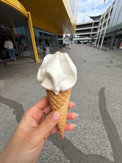 A close-up shot shows a hand holding a soft-serve ice cream cone in a waffle cone. The ice cream is white and swirled. The hand has light skin tone and a silver ring on the ring finger. The background is blurred and appears to be an outdoor public space with a large yellow building with glass doors and windows on the left. The ground is paved with grey concrete. In the distance, there is a multi-story parking garage and other buildings. The sky is visible at the top of the frame and appears partly cloudy. The image is taken during the daytime.