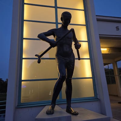 The image shows a bronze statue of a man with a broken tool or implement, possibly a sickle, held in his hands. The statue is positioned in front of a large, illuminated window composed of multiple panes. To the right of the statue, a portion of a building is visible, with the letter "S" prominently displayed. The scene is outdoors, with a twilight sky visible in the upper left corner and trees in the background on the left. The statue is on a stone pedestal. There is text on the window in the lower left, and a sign on the building's wall on the right. This sculpture is known as "The Broken Ploughman" and is located in Piešťany, Slovakia, near the Kolonádový bridge.