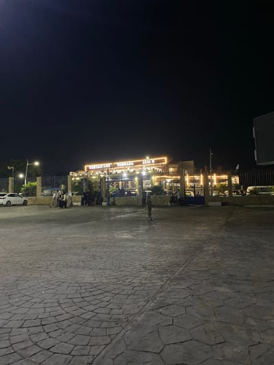 The image depicts the exterior of a restaurant named "RESTAURANT TERRAZA JANI" at night, located in Tipaza, Algeria. The establishment is illuminated by warm-toned string lights and features outdoor seating. Several people are visible, some standing in front of the restaurant and others near parked cars. A white car is parked to the left of the frame, and other vehicles are present within the restaurant's premises. The foreground is a paved area with a patterned stone surface. The sky above is dark, indicating nighttime.