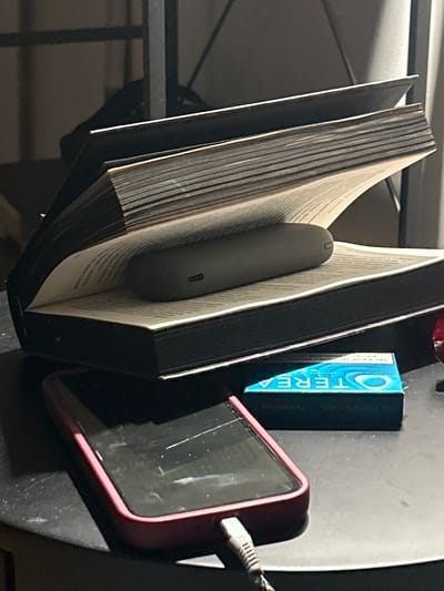 The image depicts a still life composition on a dark, textured surface, likely a table. The focal point is an open, thick book, possibly a journal or a large format novel, with its pages splayed open to reveal text. Nestled within the opened pages is a dark, elongated, oval-shaped electronic device, possibly a portable charger or a similar gadget. The device appears to have a small, circular indentation or button.
In front of the book, and slightly to the right, is a bright blue and white rectangular box. The text on the box is partially visible and reads "OTEREA" in large lettering. Below this, smaller text is visible, including "TURQUOISE". This suggests the box contains some kind of product, possibly related to oral care or vaporizers.
To the bottom left of the frame, a smartphone with a pink or red case is visible. The screen is dark, indicating it's either off or in sleep mode. A charging cable, grey with a ribbed texture, is plugged into the bottom of the phone. The cable extends out of frame to the lower right.
The background is dark and appears to be a shelf or furniture structure, with diagonal lines suggesting support beams or shelving. The lighting is dim, with a strong light source coming from the left, casting shadows and highlighting the textures of the objects. The overall impression is one of a personal space, perhaps a desk or bedside table, where technology and reading materials are present.
There is no information in the image to suggest the location in the city of Siedlce, Poland.