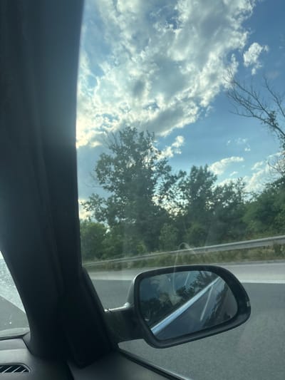 The view from inside a vehicle shows a bright sky filled with white and gray clouds. Tree branches are visible against the sky. A car's side mirror reflects a road with white lines, trees, and the sky. The interior of the vehicle is visible, including the window frame and part of the dashboard. The location in the city of Kipra, Bulgaria, cannot be determined from the provided image.